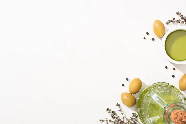 Сверху вид на оливковое масло в миске и бутылку рядом с зелеными оливками, травы и черный перец на белом фоне — стоковое фото