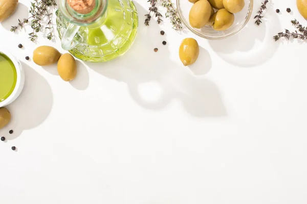 Vue du dessus de l'huile d'olive dans un bol en verre et une bouteille près des olives vertes, des herbes et du poivre noir sur fond blanc — Photo de stock