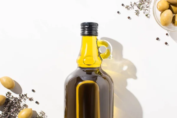 Vista dall'alto dell'olio d'oliva in bottiglia vicino alle olive verdi in ciotola, erba e pepe nero su sfondo bianco — Foto stock