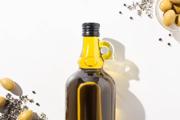 Vue du dessus de l'huile d'olive en bouteille près des olives vertes dans un bol, herbes et poivre noir sur fond blanc — Photo de stock