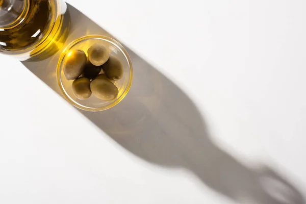 Вид зверху на оливкову олію в пляшці біля зелених оливок в мисці в тіні на білому тлі — стокове фото