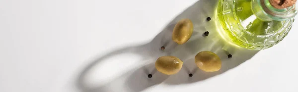 Вид зверху на оливкову олію в пляшці біля зелених оливок і чорного перцю в тіні на білому тлі, панорамна орієнтація — стокове фото