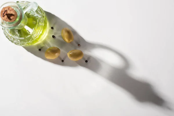 Ansicht von Olivenöl in der Flasche in der Nähe von grünen Oliven und schwarzem Pfeffer im Schatten auf weißem Hintergrund — Stockfoto