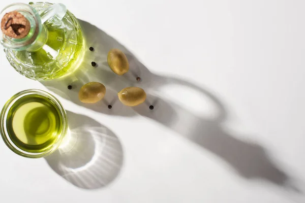 Top view of olive oil in bottle and bowl near green olives and black pepper in shadow on white background — Stock Photo