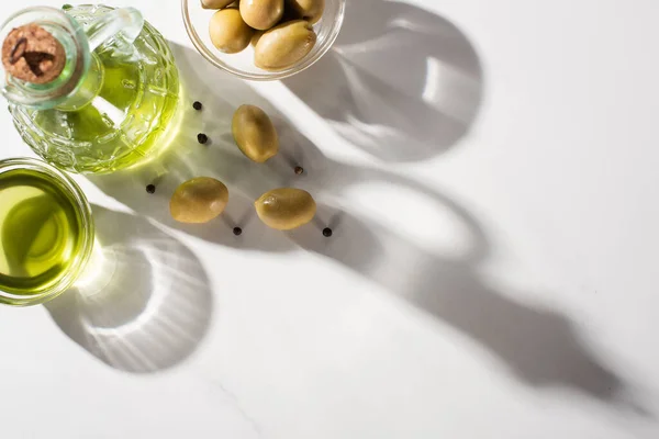 Vista superior del aceite de oliva en botella y tazón cerca de aceitunas verdes y pimienta negra en sombra sobre fondo blanco - foto de stock