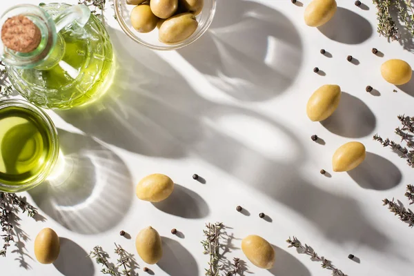 Вид зверху на оливкову олію в пляшці і мисці біля трави, зелені оливки і чорний перець на білому тлі з тіні — стокове фото