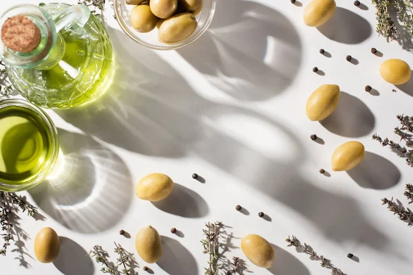 Top view of olive oil in bottle and bowl near herb, green olives and black pepper on white background with shadow — Stock Photo