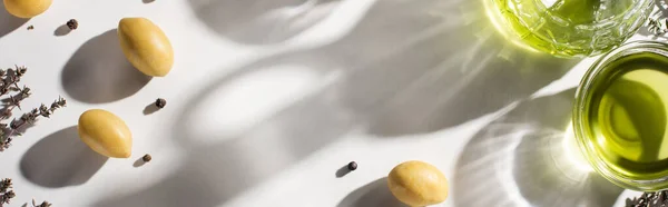 Вид на оливковое масло в бутылке и миске рядом с травой, зеленые оливки и черный перец на белом фоне с тенью, панорамная ориентация — стоковое фото
