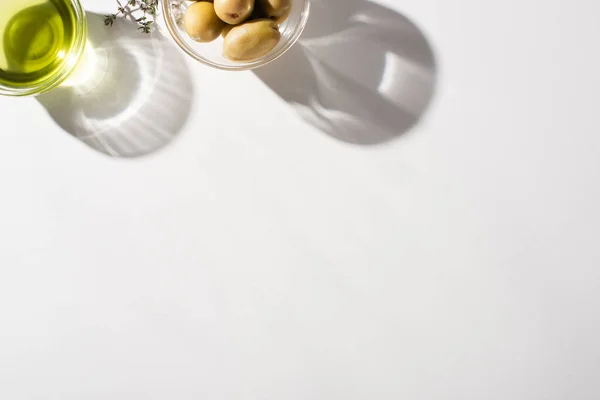 Vue du dessus de l'huile d'olive dans un bol près de l'herbe, olives vertes sur fond blanc avec ombre — Photo de stock