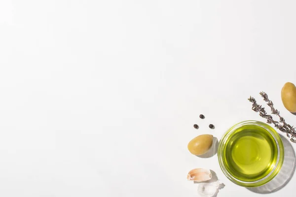 Vista dall'alto dell'olio d'oliva in ciotola vicino a olive verdi, aglio, erbe aromatiche e pepe nero su sfondo bianco — Foto stock