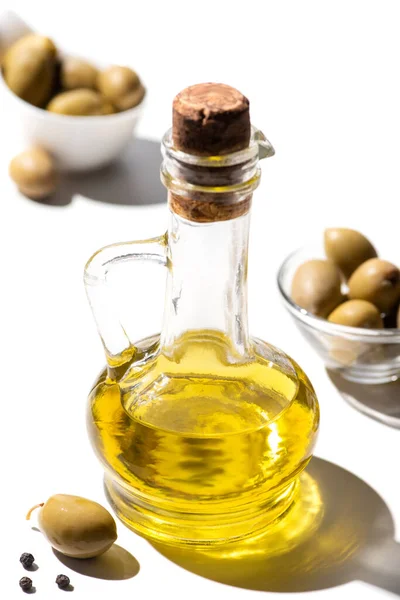 Enfoque selectivo de aceite de oliva en frasco cerca de aceitunas verdes y pimienta negra sobre fondo blanco - foto de stock