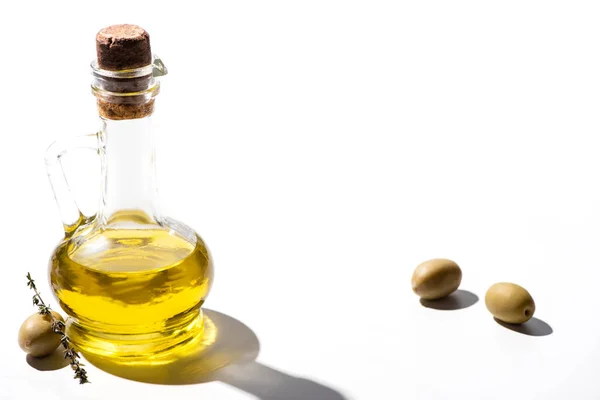Оливковое масло в банке рядом с зелеными оливками с травой на белом фоне — стоковое фото