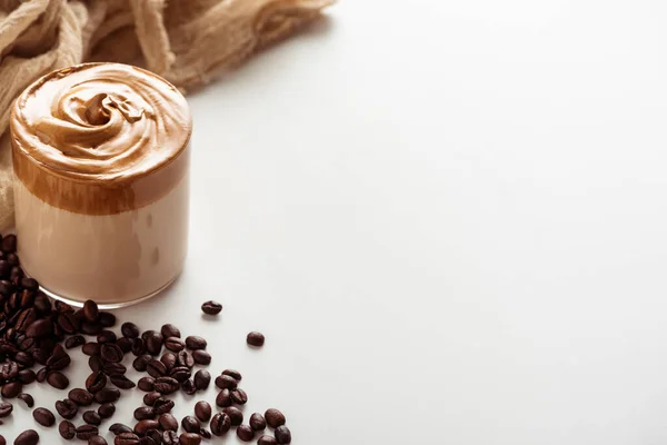 Foyer sélectif de délicieux café Dalgona en verre près de grains de café et tissu sur fond blanc — Photo de stock