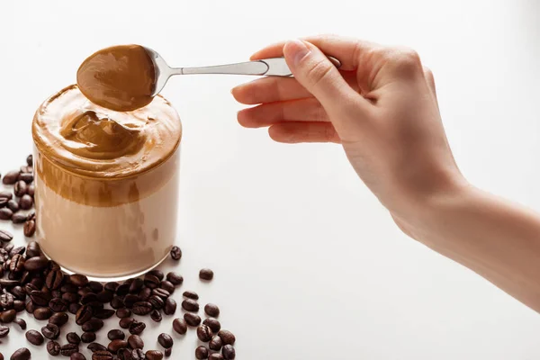 Vista ritagliata della donna che tiene cucchiaio con schiuma vicino delizioso caffè Dalgona in vetro e chicchi di caffè su sfondo bianco — Foto stock