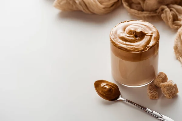 Delicioso café Dalgona em vidro perto de pano, açúcar mascavo e colher no fundo branco — Fotografia de Stock