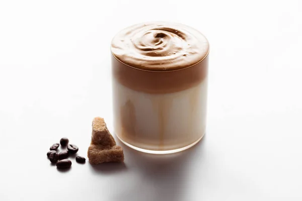 Délicieux café Dalgona en verre près de grains de café et de sucre granulé brun sur fond blanc — Photo de stock