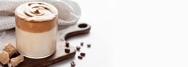 Selective focus of delicious Dalgona coffee in glass near coffee beans, brown sugar on cutting board and napkin on white background, panoramic shot — Stock Photo