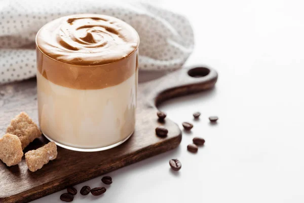 Selektiver Fokus von köstlichem Dalgona-Kaffee im Glas in der Nähe von Kaffeebohnen, braunem Zucker auf Schneidebrett und Serviette auf weißem Hintergrund — Stockfoto