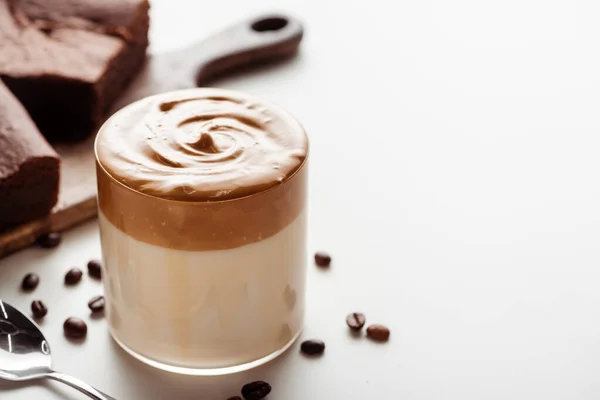 Selektiver Fokus von köstlichem Dalgona-Kaffee im Glas in der Nähe von Kaffeebohnen, Schneidebrett und Löffel auf weißem Hintergrund — Stockfoto