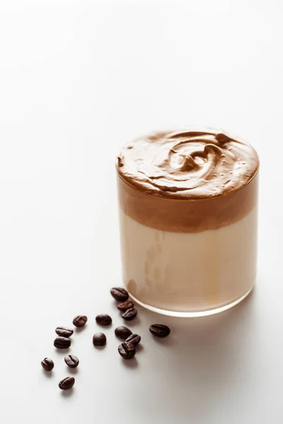 Вкусный кофе Dalgona в стекле возле кофейных зерен на белом фоне — стоковое фото