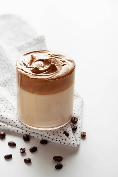 Delicioso café Dalgona em vidro perto de grãos de café e guardanapo no fundo branco — Fotografia de Stock
