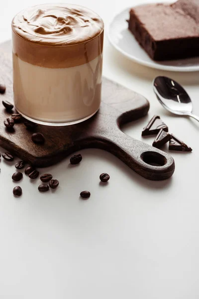 Enfoque selectivo de delicioso café Dalgona en vidrio sobre tabla de cortar de madera cerca de granos de café, chocolate y cuchara sobre fondo blanco - foto de stock