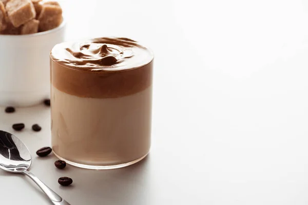 Foco seletivo de delicioso café Dalgona em vidro perto de colher, grãos de café e açúcar mascavo no fundo branco — Fotografia de Stock