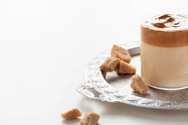 Köstlicher Dalgona Kaffee im Glas in der Nähe von braunem Zucker auf silbernem Tablett auf weißem Hintergrund — Stockfoto