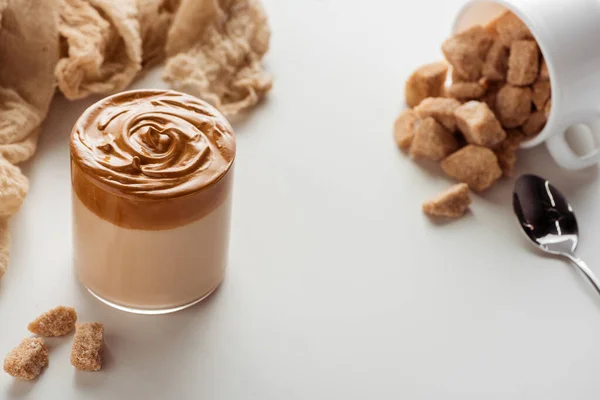 Selektiver Fokus von köstlichem Dalgona-Kaffee im Glas in der Nähe von Löffel, Tasse, Tuch und braunem Kristallzucker auf weißem Hintergrund — Stockfoto