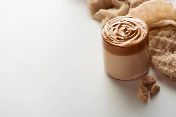 Delicioso café Dalgona em vidro perto de pano e açúcar mascavo granulado no fundo branco — Fotografia de Stock