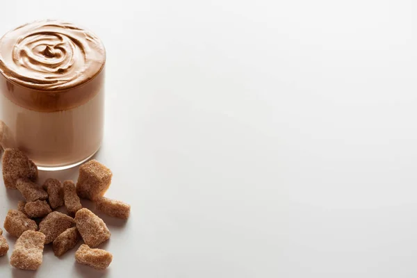 Köstlicher Dalgona-Kaffee im Glas in der Nähe von braunem Zucker auf weißem Hintergrund — Stockfoto