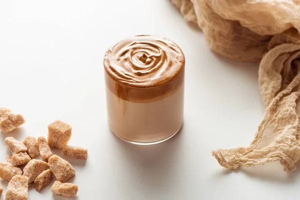 Delicioso café Dalgona en vidrio cerca de tela y azúcar morena granulada sobre fondo blanco - foto de stock