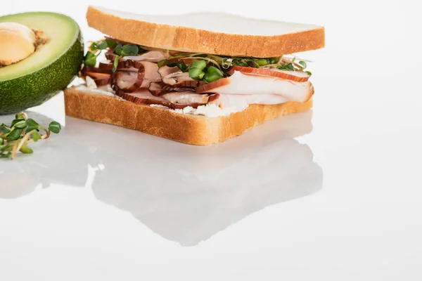 Свежий вкусный сэндвич с мясом и ростками на белой поверхности возле авокадо — стоковое фото