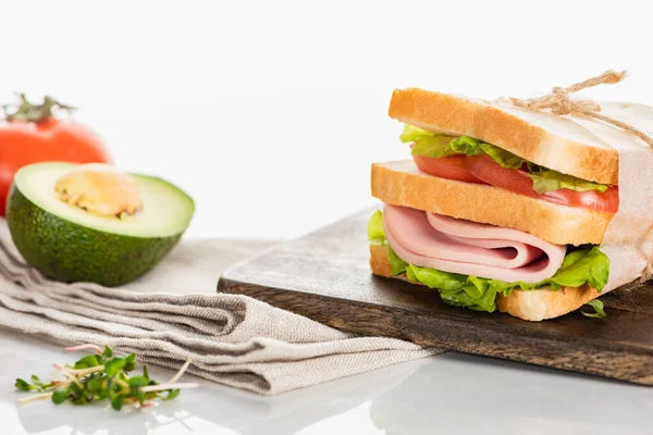 Delicioso sándwich fresco con salchicha en rodajas y lechuga en tabla de cortar de madera cerca de la servilleta y el aguacate en la superficie blanca — Stock Photo