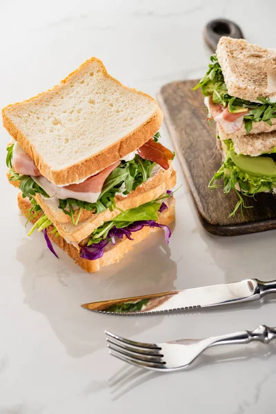 Fresh sandwiches with meat on marble white surface with cutlery and wooden cutting board — Stock Photo