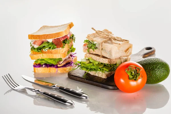 Bocadillos frescos en la tabla de cortar de madera cerca de cubiertos y aguacate, tomate en la superficie blanca - foto de stock