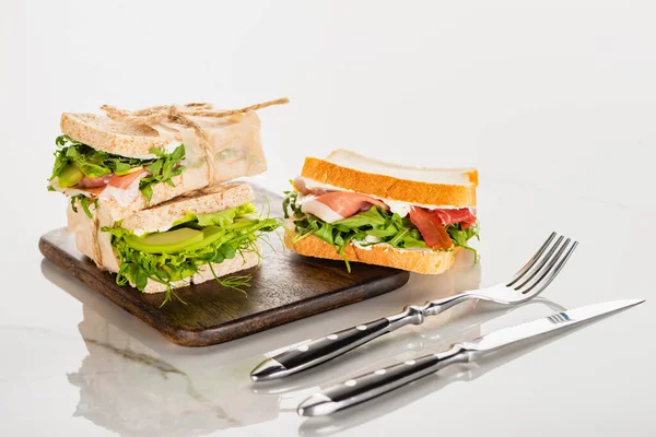 Свежие бутерброды на деревянной доске возле столовых приборов на поверхности белого мрамора — стоковое фото