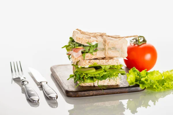 Frische grüne Sandwiches auf hölzernem Schneidebrett in der Nähe von Besteck und Tomaten auf weißer Oberfläche — Stockfoto