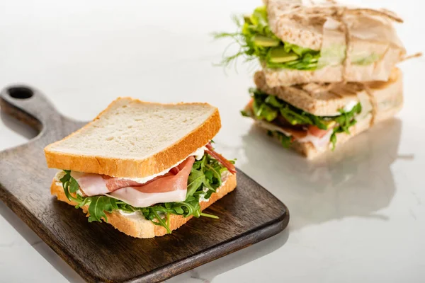 Selective focus of fresh green sandwich with prosciutto on wooden cutting board on white marble surface — Stock Photo