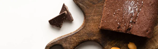 Vista superior de deliciosa peça de brownie na placa de corte de madeira com chocolate no fundo branco, tiro panorâmico — Fotografia de Stock