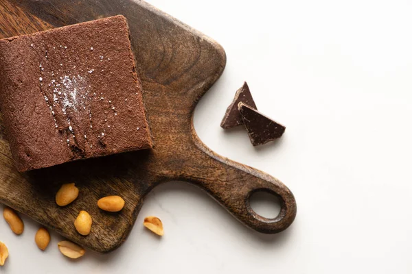 Vue du dessus de délicieux morceau de brownie sur planche à découper en bois avec des noix et du chocolat sur fond blanc — Photo de stock