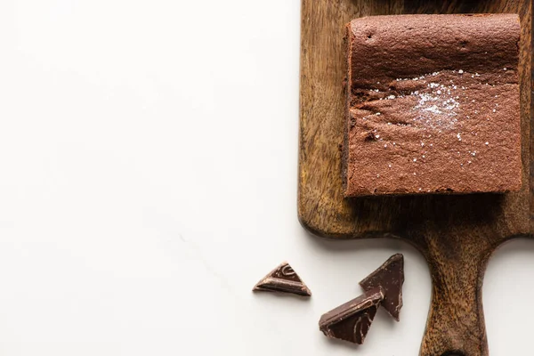 Draufsicht auf leckeres Brownie-Stück auf Holzschneidebrett mit Schokolade auf weißem Hintergrund — Stockfoto