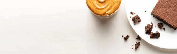 Top view of delicious brownie piece on plate near peanut butter on white background, panoramic shot — Stock Photo