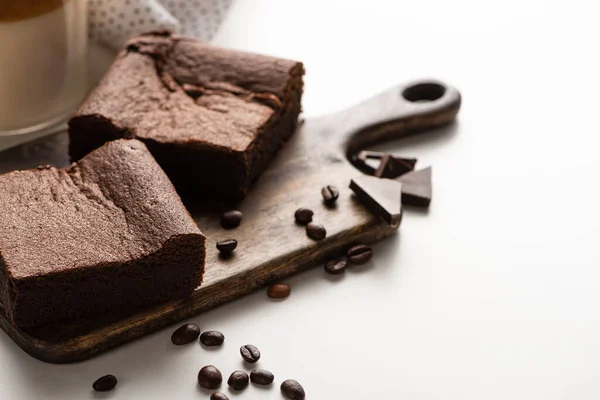 Délicieux morceaux de brownie sur planche à découper en bois avec grains de café et chocolat sur fond blanc — Photo de stock
