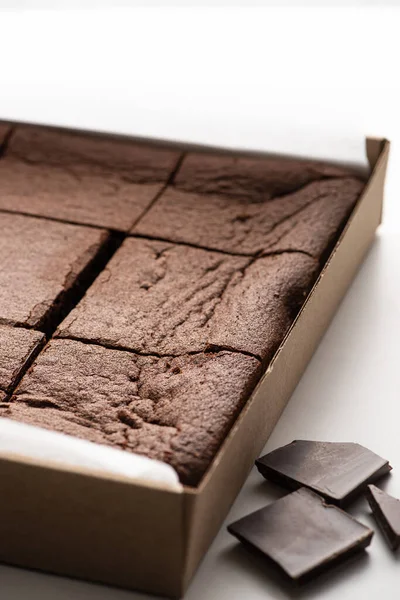 Vista da vicino di deliziosi pezzi di brownie in scatola di cartone vicino al cioccolato fondente su sfondo bianco — Foto stock