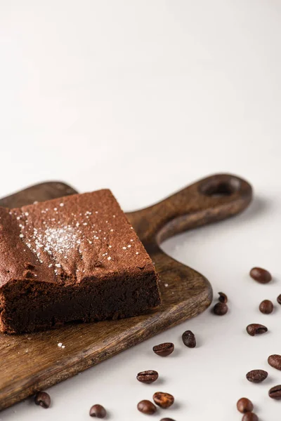 Delizioso pezzo brownie su tagliere in legno con chicchi di caffè su sfondo bianco — Foto stock