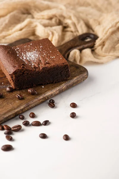 Delicioso pedaço de brownie na placa de corte de madeira com grãos de café perto de pano no fundo branco — Fotografia de Stock