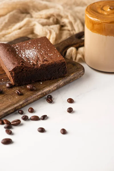 Delizioso pezzo di brownie su tagliere in legno con chicchi di caffè, caffè e stoffa su sfondo bianco — Foto stock