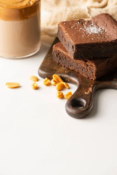 Selektiver Fokus von leckeren Brownie-Stücken auf Holzschneidebrett mit Erdnüssen in der Nähe von Kaffee im Glas auf weißem Hintergrund — Stockfoto