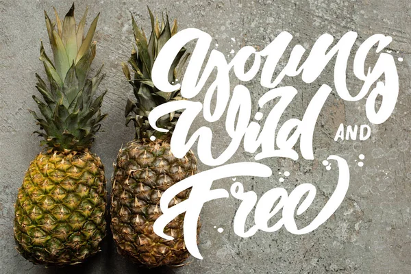 Top view of ripe pineapples on grey concrete surface with young, wild and free illustration — Stock Photo